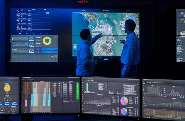 Two people observe a large screen showing the San Francisco bay area. Around them are several other screens displaying simulation data.