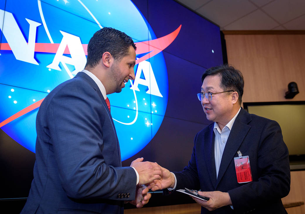 Two men wearing suits shake hands in front of a screen showing the NASA logo, which fills the wall behind them.