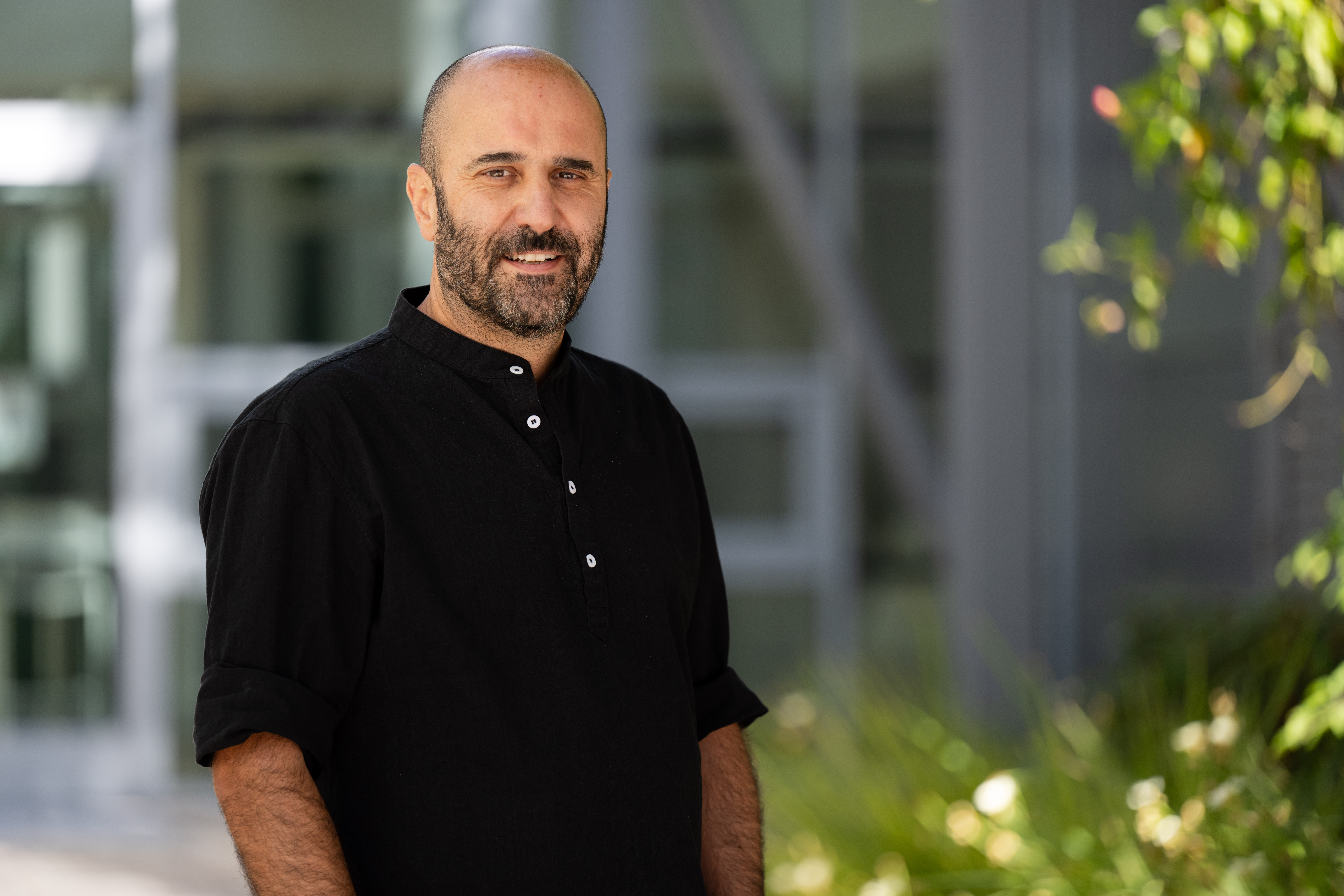 Alfonso Davila, a Hispanic person, stands in front of a building that is softly out of focus. He wears a black shirt with rolled up sleeves.