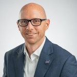 Portrait of Ian Brosnan in a blue blazer and white shirt and in glasses