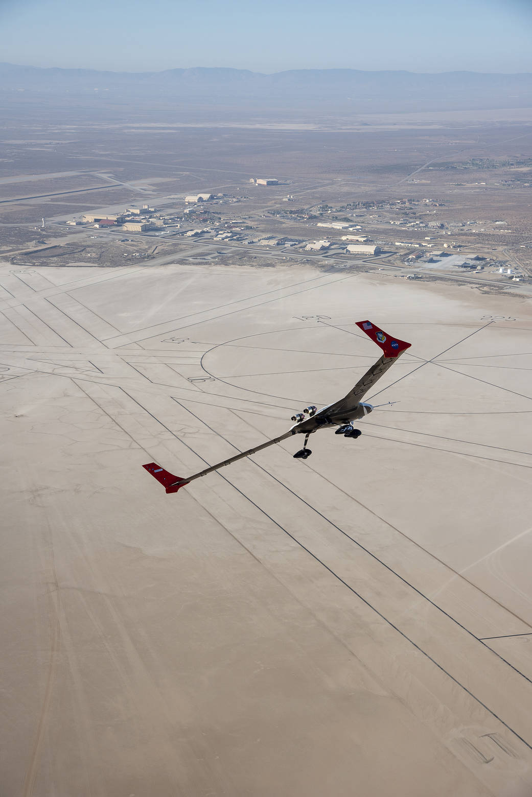 The X-56A flies over Rogers Dry Lakebed. A huge compass rose painted onto the lakebed can be seen below. Lines to the left and below the compass rose are runways.