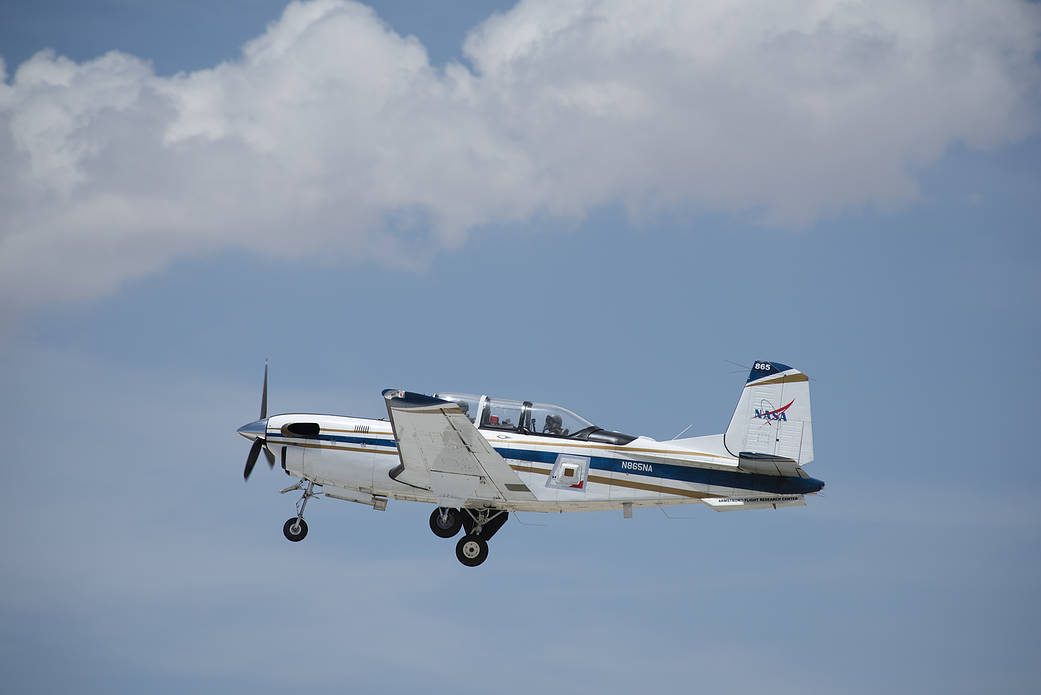 Small two-seat aircraft in flight against a blue sky with white clouds.