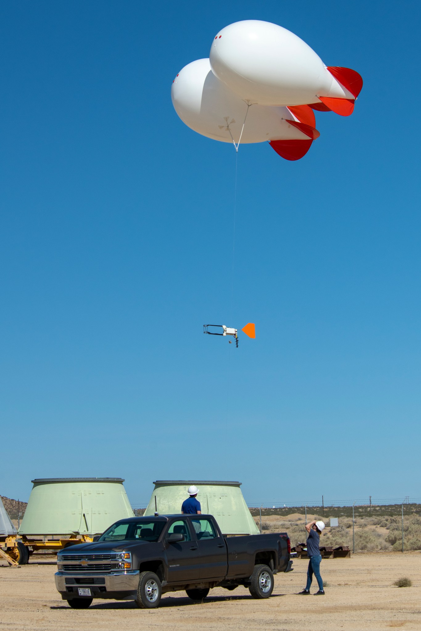 A weather instrument is attached to two balloons above it. The balloons are tied to a dark gray truck parked below.