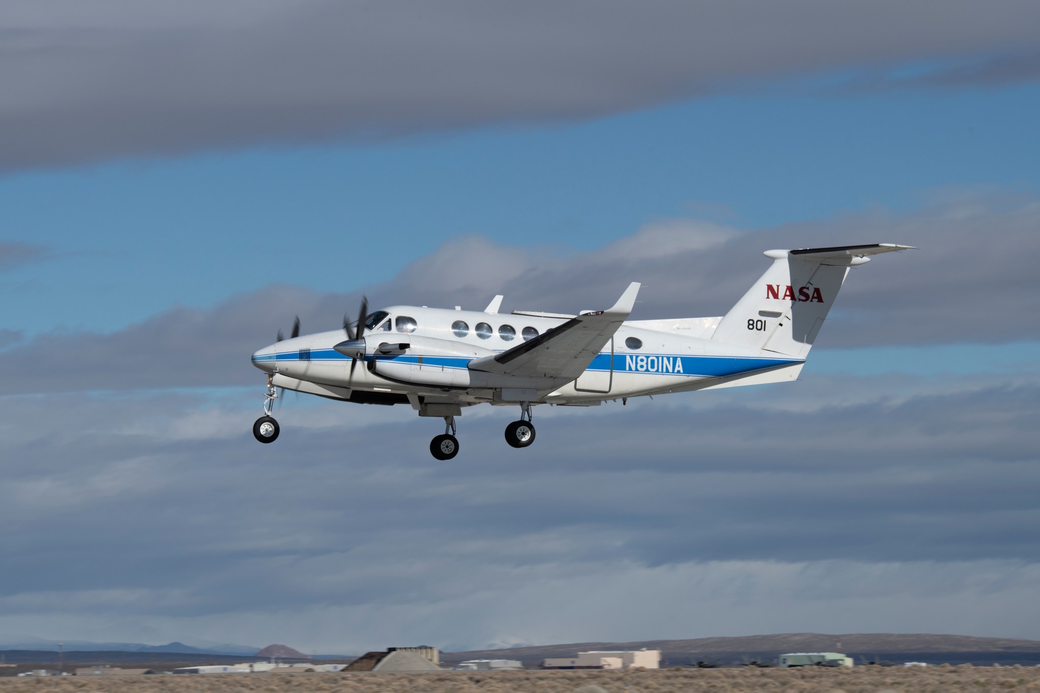 Aircraft in low flight with landing gear down. A desert town and cloudy blue sky seen in the background.