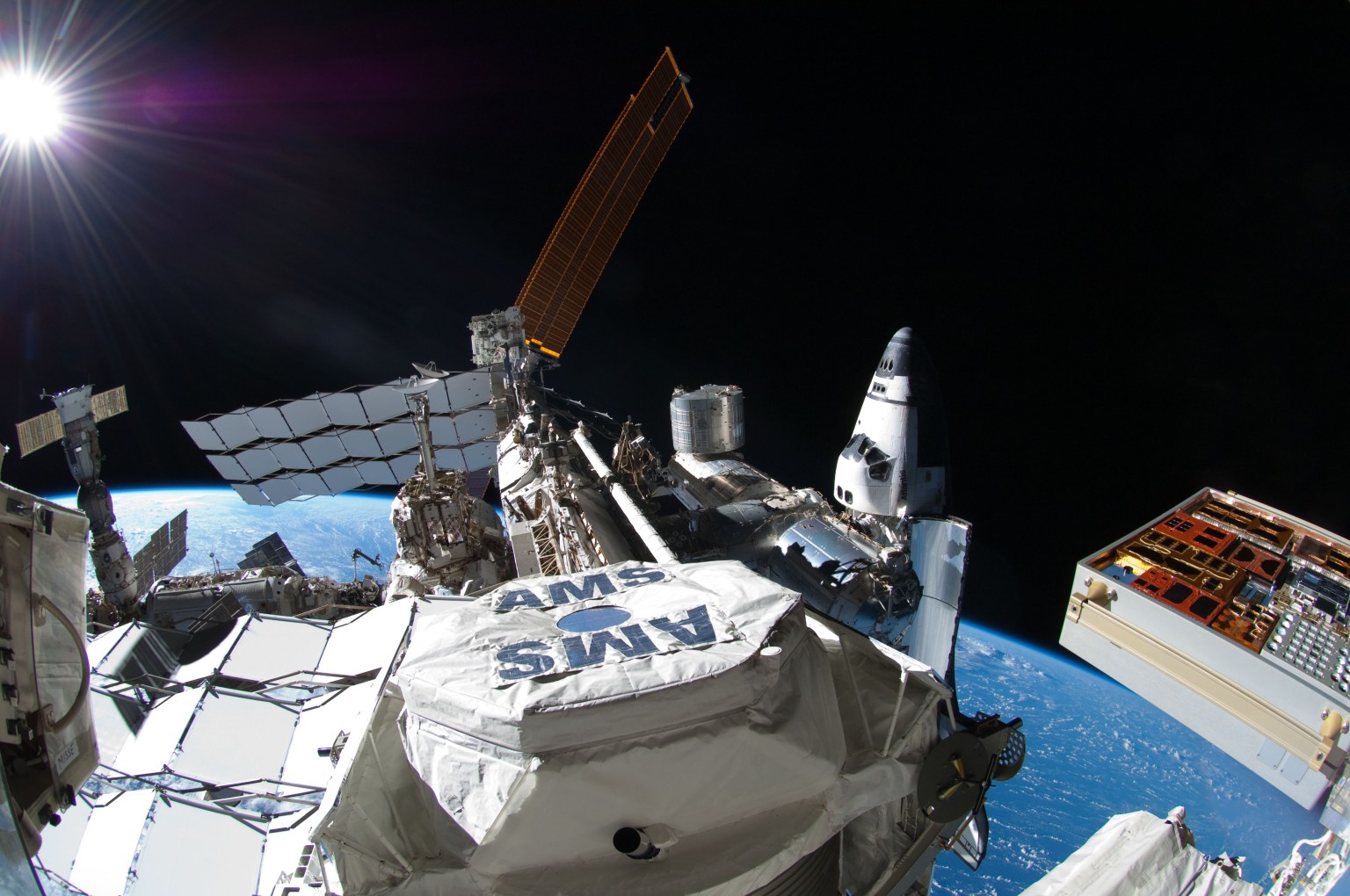 This picture, photographed by NASA astronaut Ron Garan during the spacewalk conducted on July 12, 2011, shows the International Space Station with space shuttle Atlantis docked at right and a Russian Soyuz docked to Pirs, below the sun at far left. In the center foreground is the Alpha Magnetic Spectrometer (AMS) experiment installed during the STS-134 mission. AMS is a state-of-the-art particle physics detector designed to use the unique environment of space to advance knowledge of the universe and lead to the understanding of the universe's origin by searching for antimatter and dark matter, and measuring cosmic rays.