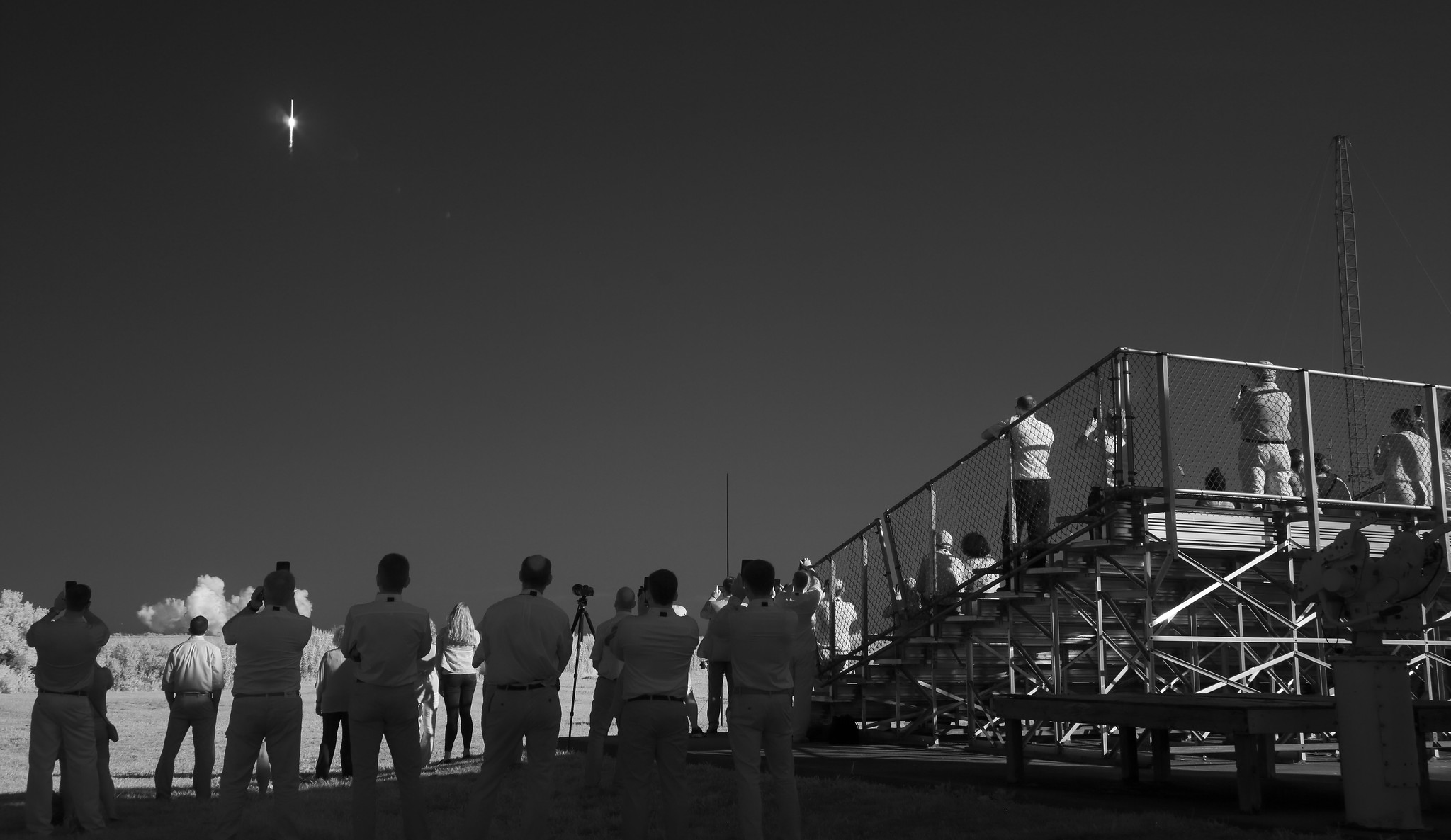 A black and white photo of bleachers with people watching a rocket launching in the background.