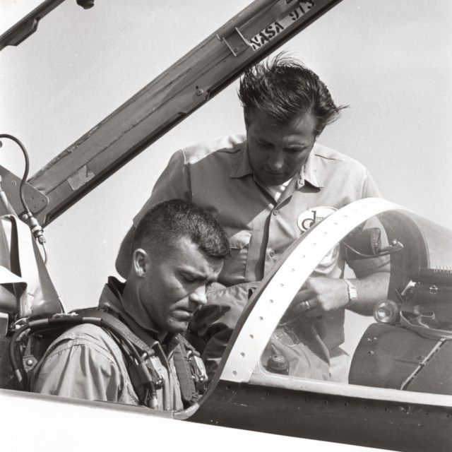 Pilot sitting in cockpit being assisted by technician.