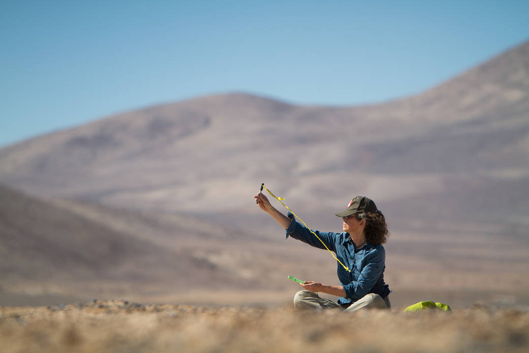 A seated figure in the desert holds a cord up in the wind to find the wind speed.