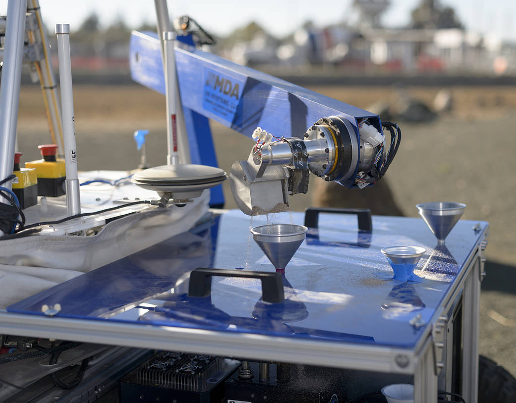 Soil collected by the ARADS rover is poured by a scoop into metal funnels on the rover’s top platform,