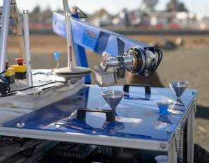 Soil collected by the ARADS rover is poured by a scoop into metal funnels on the rover’s top platform,
