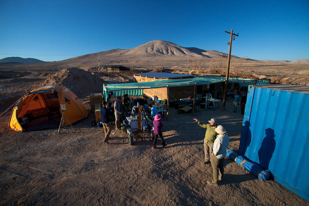 The ARADS base camp in the Atacama Desert. There are tents, the rover, and scientists talking, with mountains behind them.