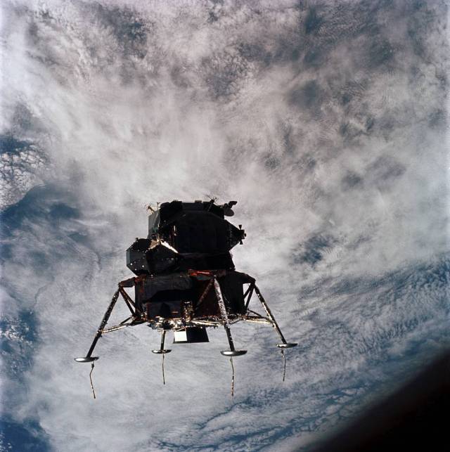 Lunar Module in orbit around the Earth. It appears almost in silhouette with clouds appearing in the background over Earth's surface.