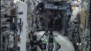 Inside the International Space Station, two astronauts work alongside two floating, cube-shaped robots