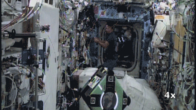 Inside the International Space Station, two astronauts work alongside two floating, cube-shaped robots