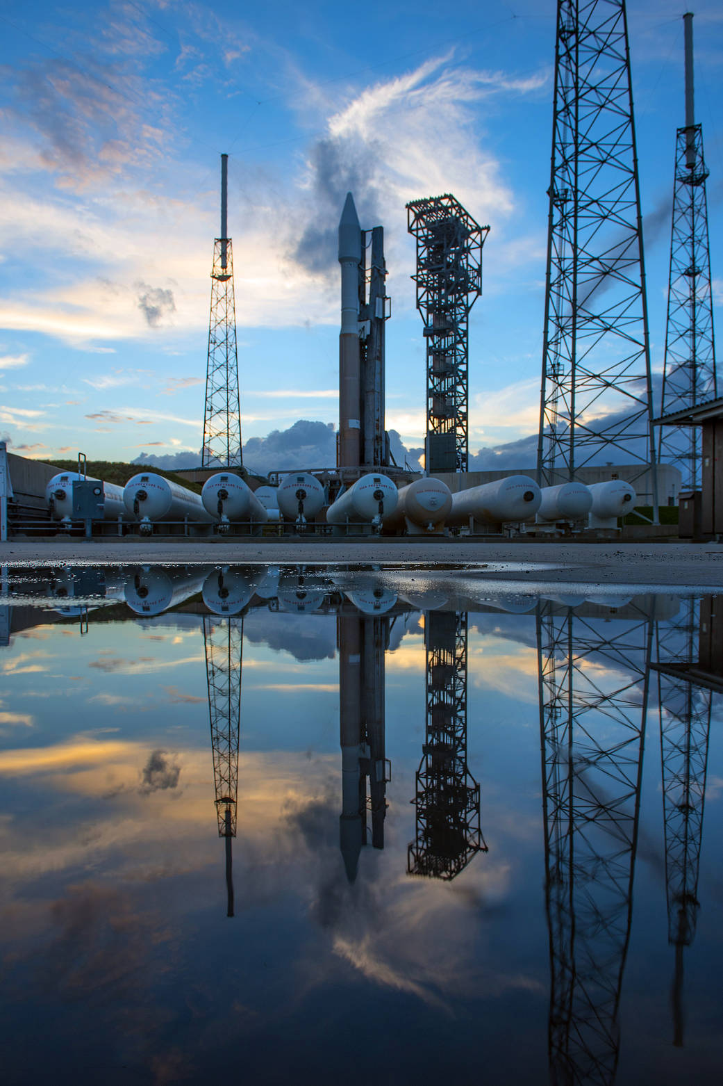 ULA Atlas V rocket with Cygnus rocket aboard at launch pad, reflected in pool of water below