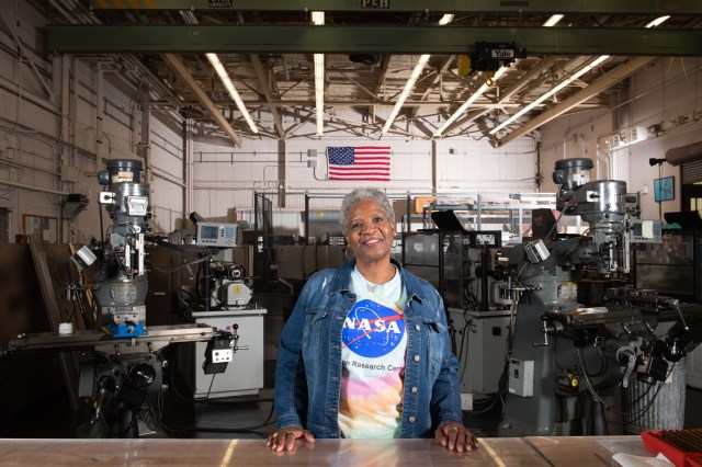 A woman faces the camera smiling. She stands in a machine shop. She wears a tie-dye NASA Glenn shirt. A large American flag hangs on the wall in the background.