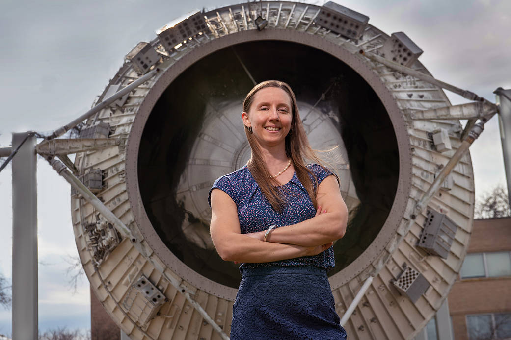 Woman stands with arms folded in front of a rocket display outdoors. 