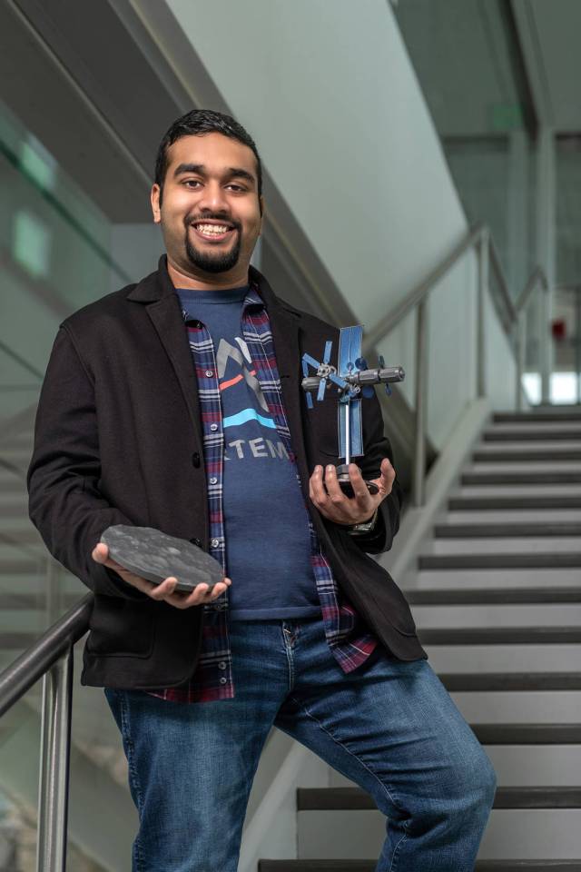 A young man smiles at camera holding a 3D printed model in one hand and the model of spacecraft in the other hand.