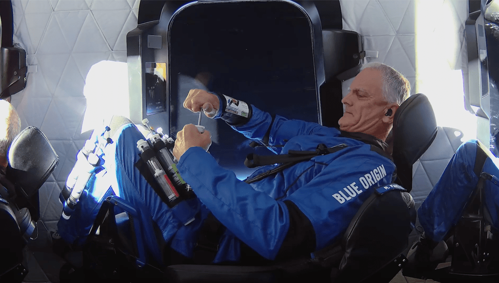 Researcher strapped into seat depresses plunger on tube. Window view is the darkness of space.