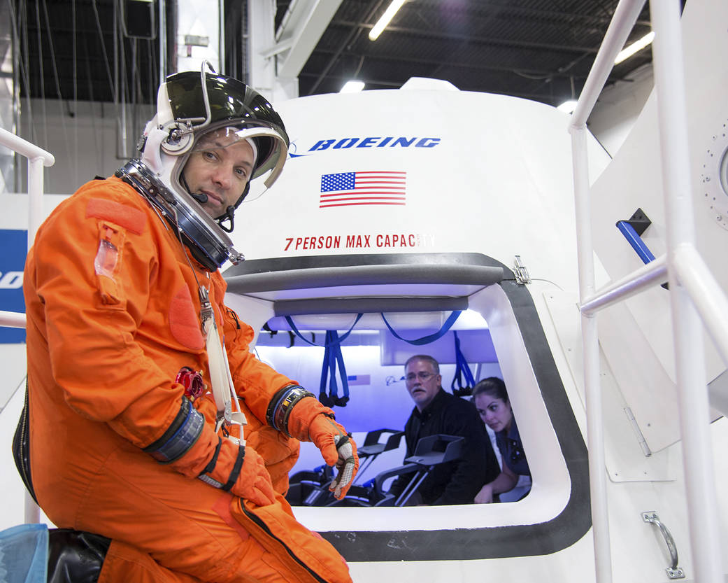 Astronaut in orange flight suit with helmet visor open outside entrance hatch of Boeing space capsule