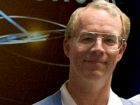 A man with light hair and glasses smiles at the camera. He wears a white collared shirt and blue jacket. Behind him, a graphic depicts the orbits of the solar system in light yellow circles against a dark red background.