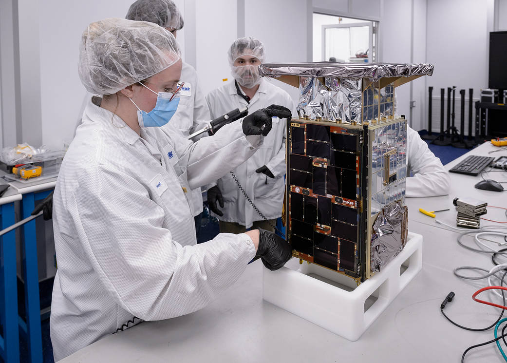 An engineer in a white lab suit holding a tape ruler to a small, microwave-sized, rectangular small satellite. One side has black panels, and others have silver panels and foil. Three team members are seen in the back.