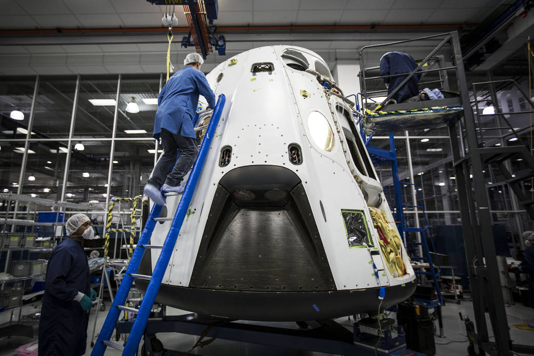 SpaceX Dragon capsule inside facility with technician climbing ladder to top and second technician standing nearby