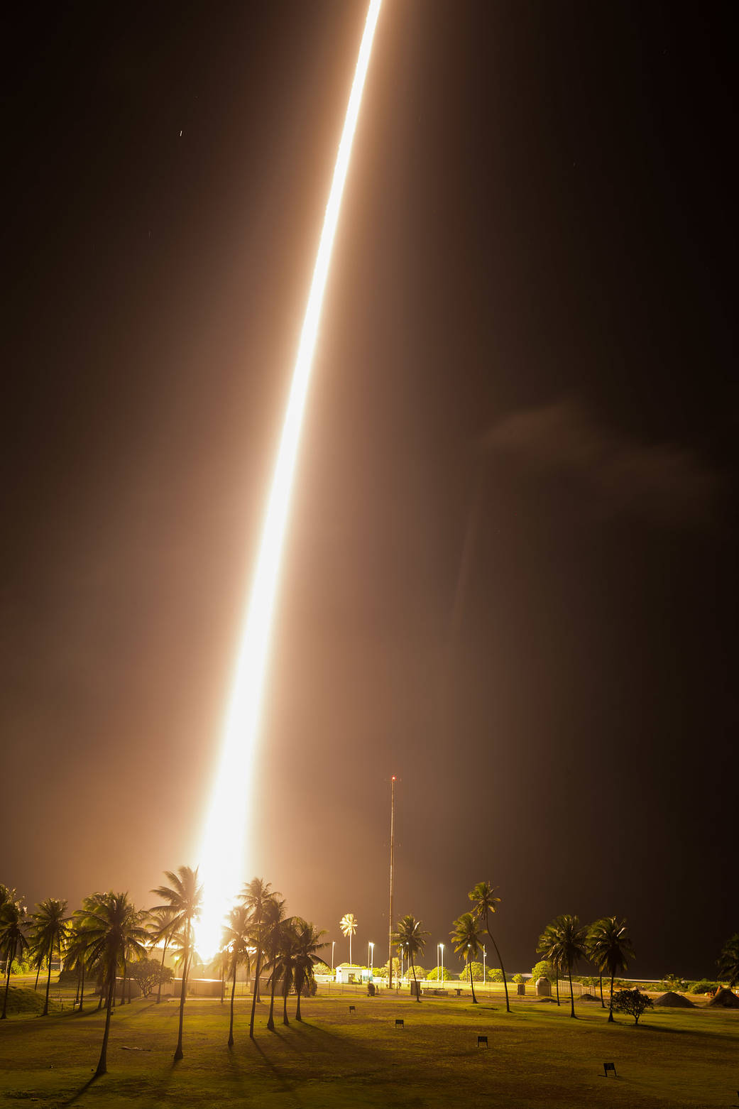 A long-exposure photo of a sounding rocket represented by a white streak, taken during the night with the landscape of green grass and palm trees lit up.