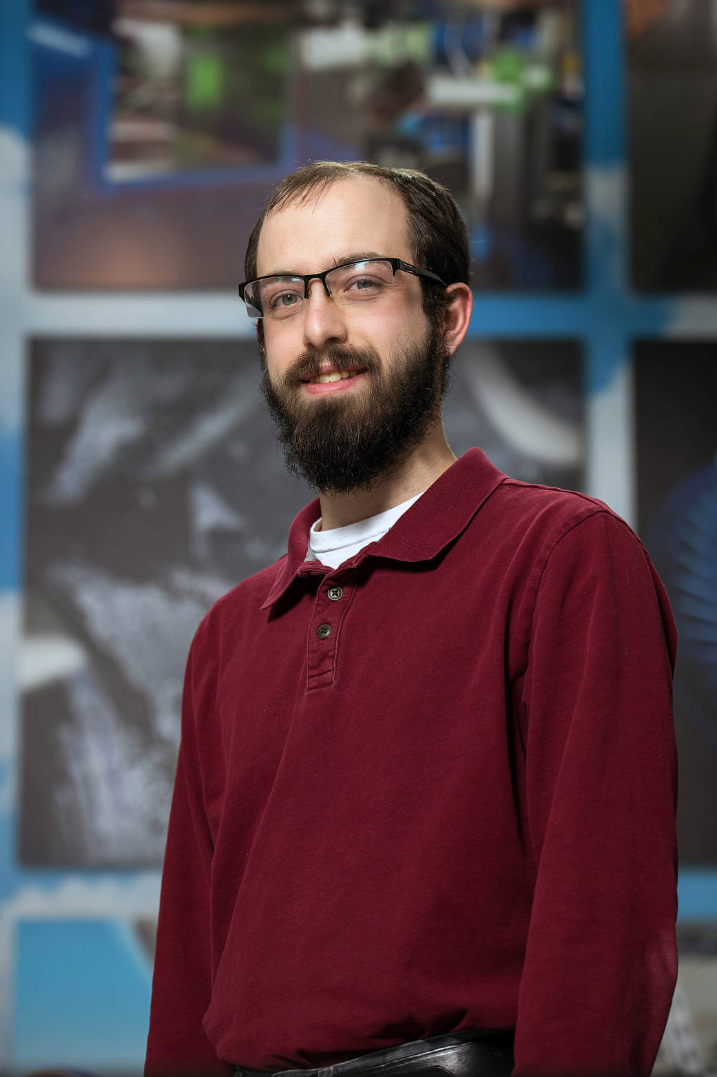 Young engineer wearing glasses and burgundy shirt smiles at camera in front of blurred background.