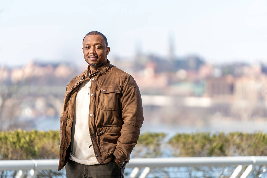 Clifford Timpson smiling and wearing a brown leather jacket while standing in front of a river with the Arlington, Virginia city-scape blurred behind him. 