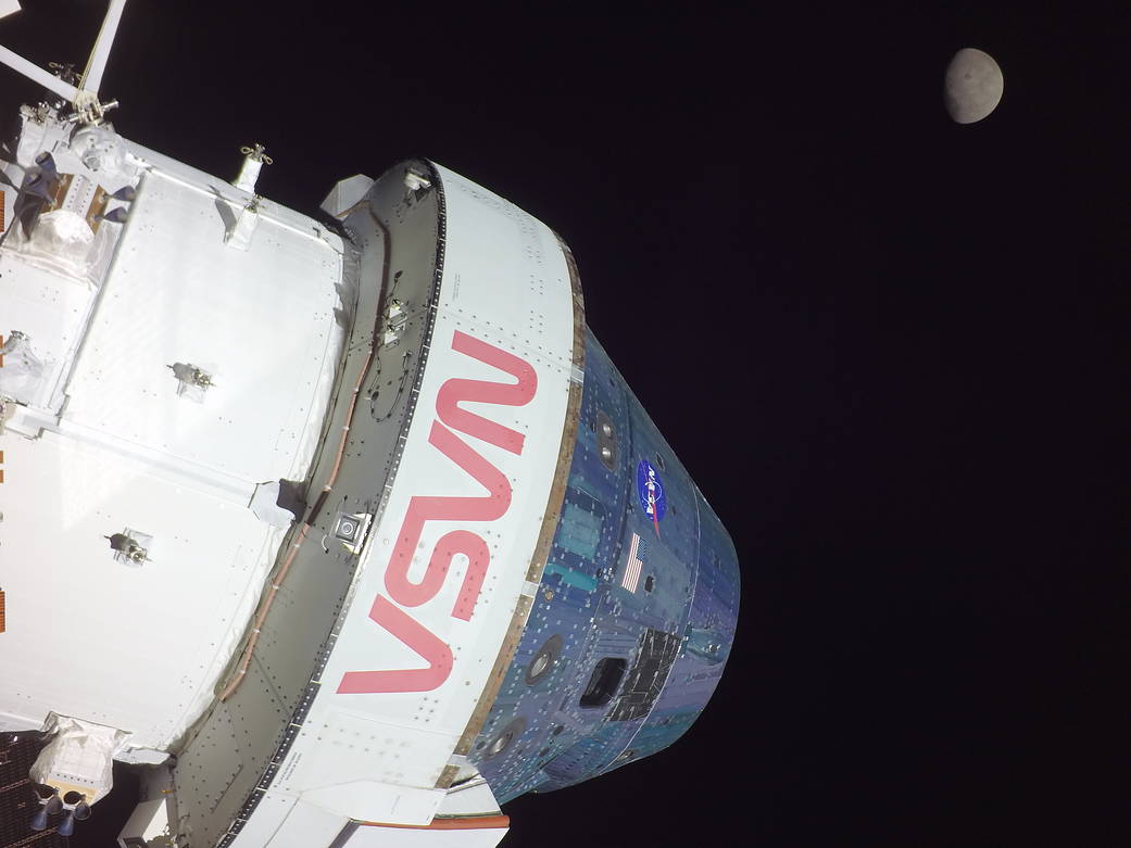 The Orion spacecraft dominates the foreground of this photo, with the NASA worm logo prominently visible in bold, red letters. In the background, the Moon appears, small and gray.