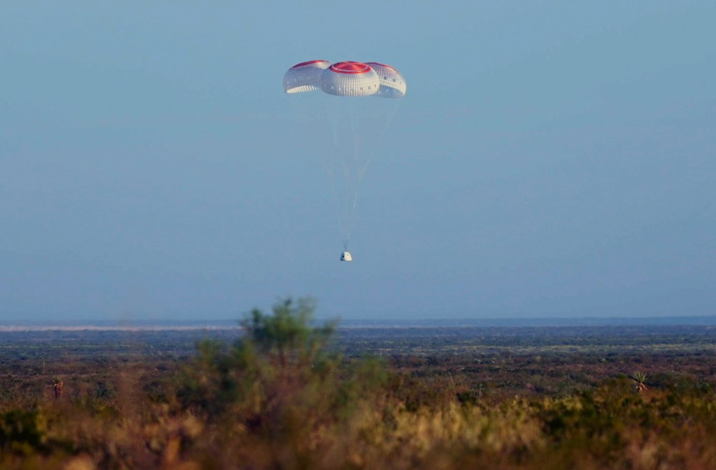 A capsule descends to the desert floor tethered to three parachutes.