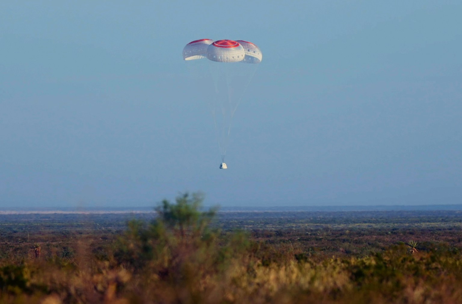 A capsule descends to the desert floor tethered to three parachutes.
