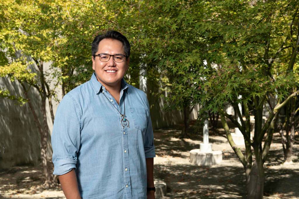 Aaron Yazzie dressed in a light blue button down shirt with his arms by his side smiling face forward at the camera with trees behind him. He's wearing black rimmed glasses and a Native American necklace. 