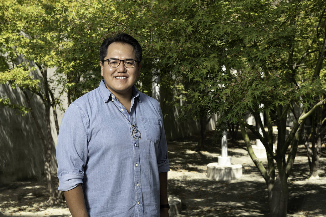Aaron Yazzie dressed in a light blue button down shirt with his arms by his side smiling face forward at the camera with trees behind him. He's wearing black rimmed glasses and a Native American necklace. 