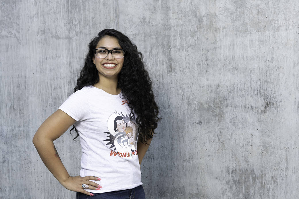 Lauren Denson wearing a white "women in space" t-shirt smiling with her hands on her hips in front of a grey wall. She's wearing black glasses and a Native American ring. 