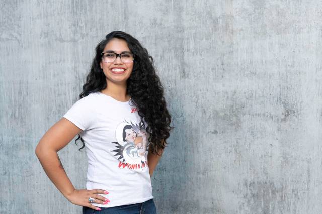 Lauren Denson wearing a white "women in space" t-shirt smiling with her hands on her hips in front of a grey wall. She's wearing black glasses and a Native American ring. 