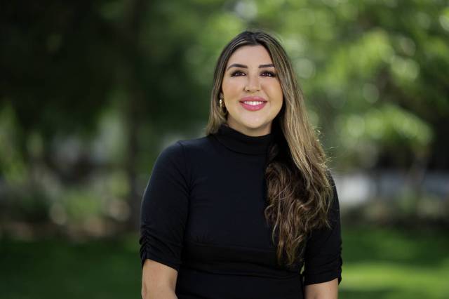 Linda Rivera wearing a black turtle neck sweater in front of green trees. 