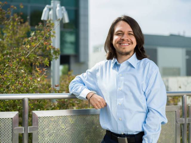 A man from Colombia with shoulder length brown hair and facial hair, smiles widely at the camera wearing a light blue button up shirt and dark pants. His right elbow props him up on the railing outside at NASA's Jet Propulsion Laboratory. 