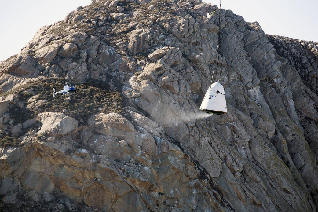 SpaceX capsule descends to land in front of rocky terrain with helicopter nearby