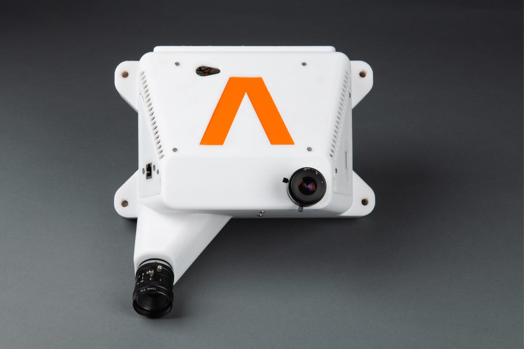 White trapezoid-shaped device with one lens extending off the lower left corner and a second lens at the lower right corner. An inverted orange “V” is on the front.