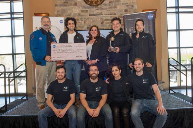 Members of AI. SpaceFactory receive a large check as an award for winning the final round of NASA's 3D Printed Habitat Challenge. Five people stand in the back and four people sit in the front on a stage, with the check being held by a man in a blue NASA jacket on the far left and a member of AI. SpaceFactory holding the check next to the man in the blue jacket. All nine people smile brightly.