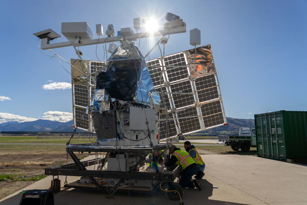 The SuperBIT payload on a stand being worked on from underneath by technicians. The payload is made up a multiple solar panels, a bar across the top with various instrumentation, and many electronic components make up the body of the payload.