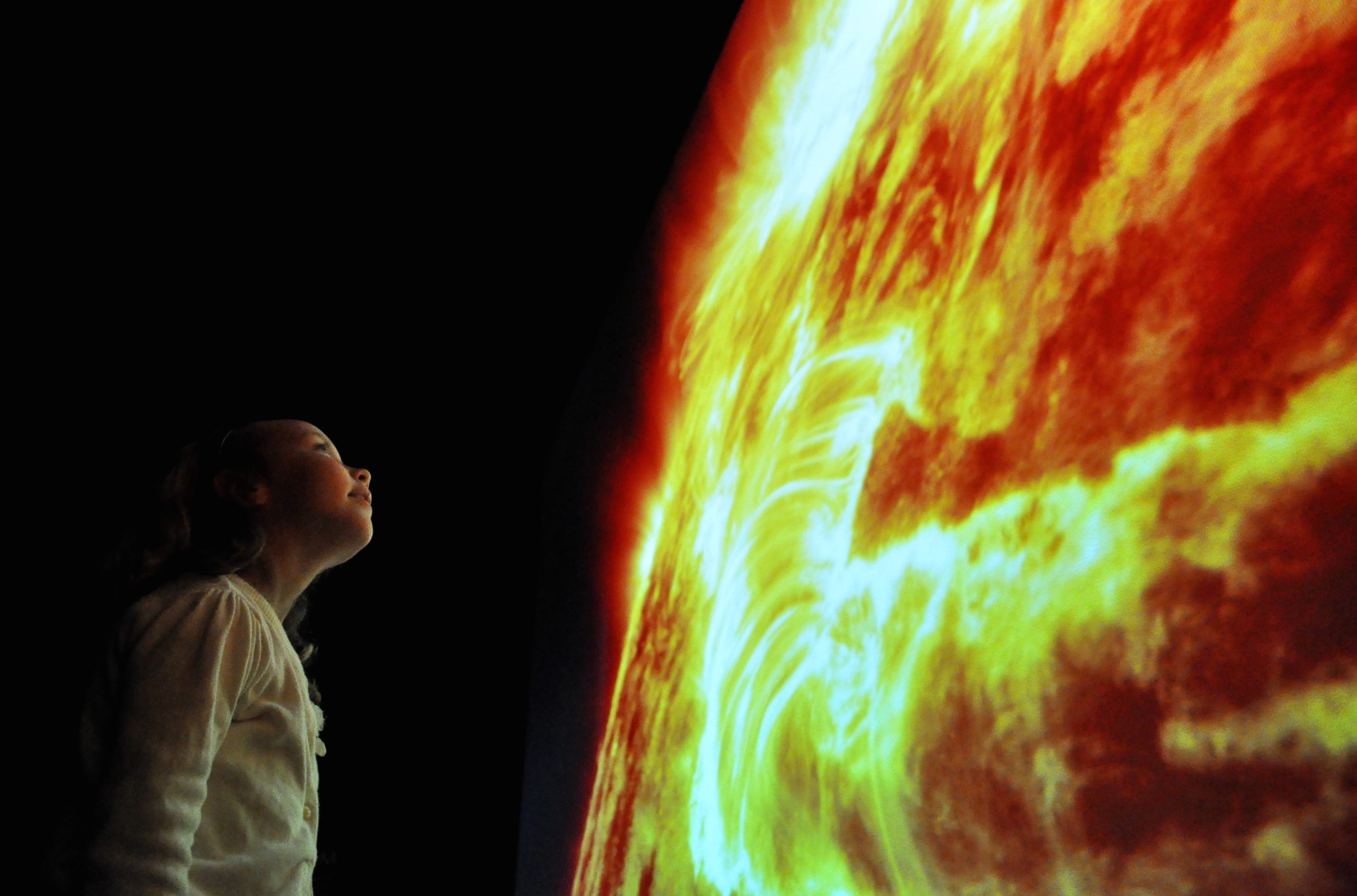 A young girl stands in front of a huge, glowing image of the Sun, appearing as a massive red and yellow swirling surface covering the whole right side of the image. The room is completely dark except for the light shining off the screen, illuminating the girl's face in bright contrasts. She wears a white sweater and looks upward at the Sun.