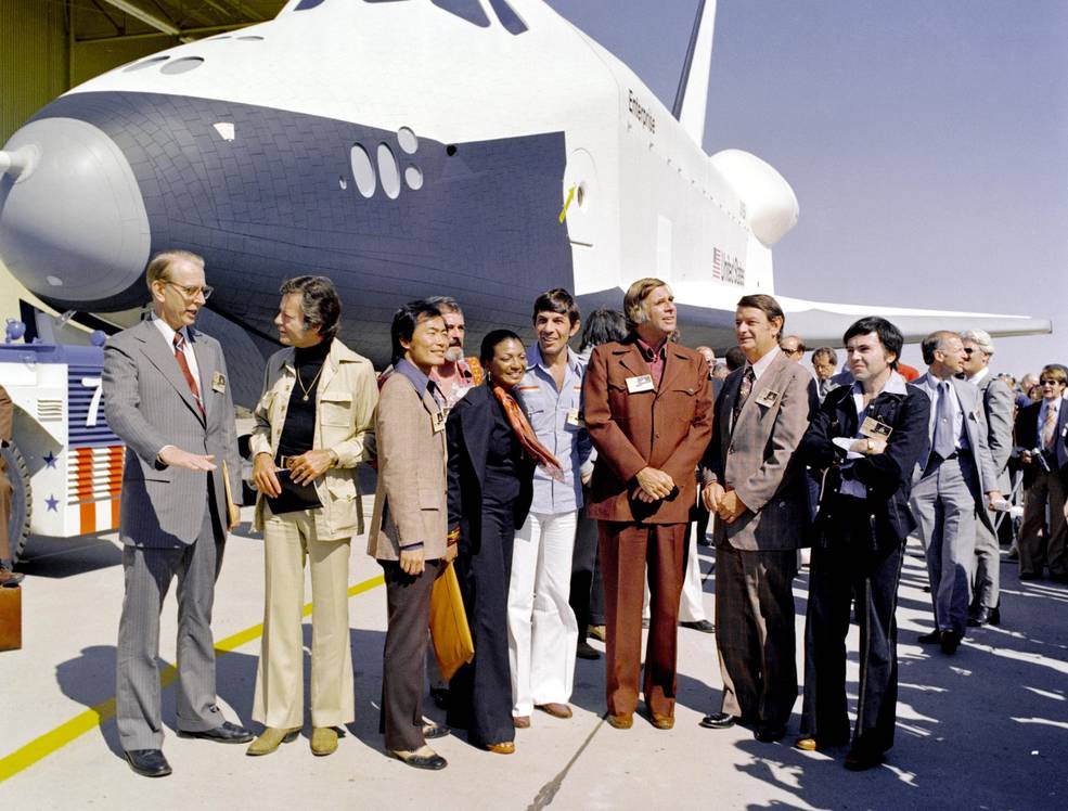 a space shuttle orbiter named Enterprise sits behind NASA officials and actors from the TV series Star Trek.