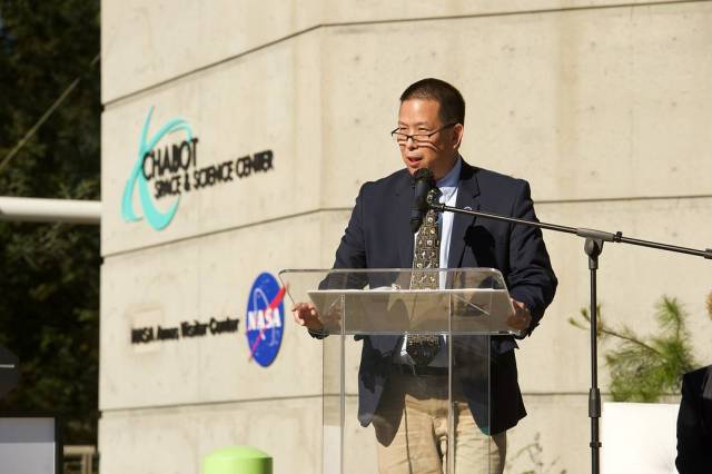 Eugene Tu speaking in front of a podium, with a building in the background labelled Chabot Space and Science Center and NASA Ames Visitor Center, with the NASA logo.
