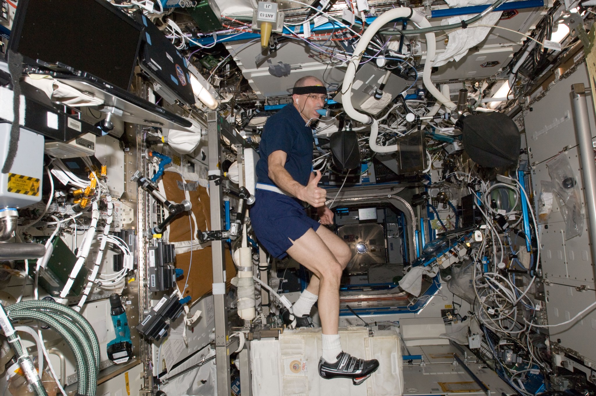 Pettit is wearing a blue t-shirt and shorts, earbuds, a head band, and black running shoes, giving a thumbs up from the station’s cycling machine, a white suitcase-sized box with bicycle pedals. Pettit holds a blue mouthpiece in his mouth attached to a tube that measures his oxygen uptake. The walls of the station around him are covered with equipment, laptops, hoses, and cords.