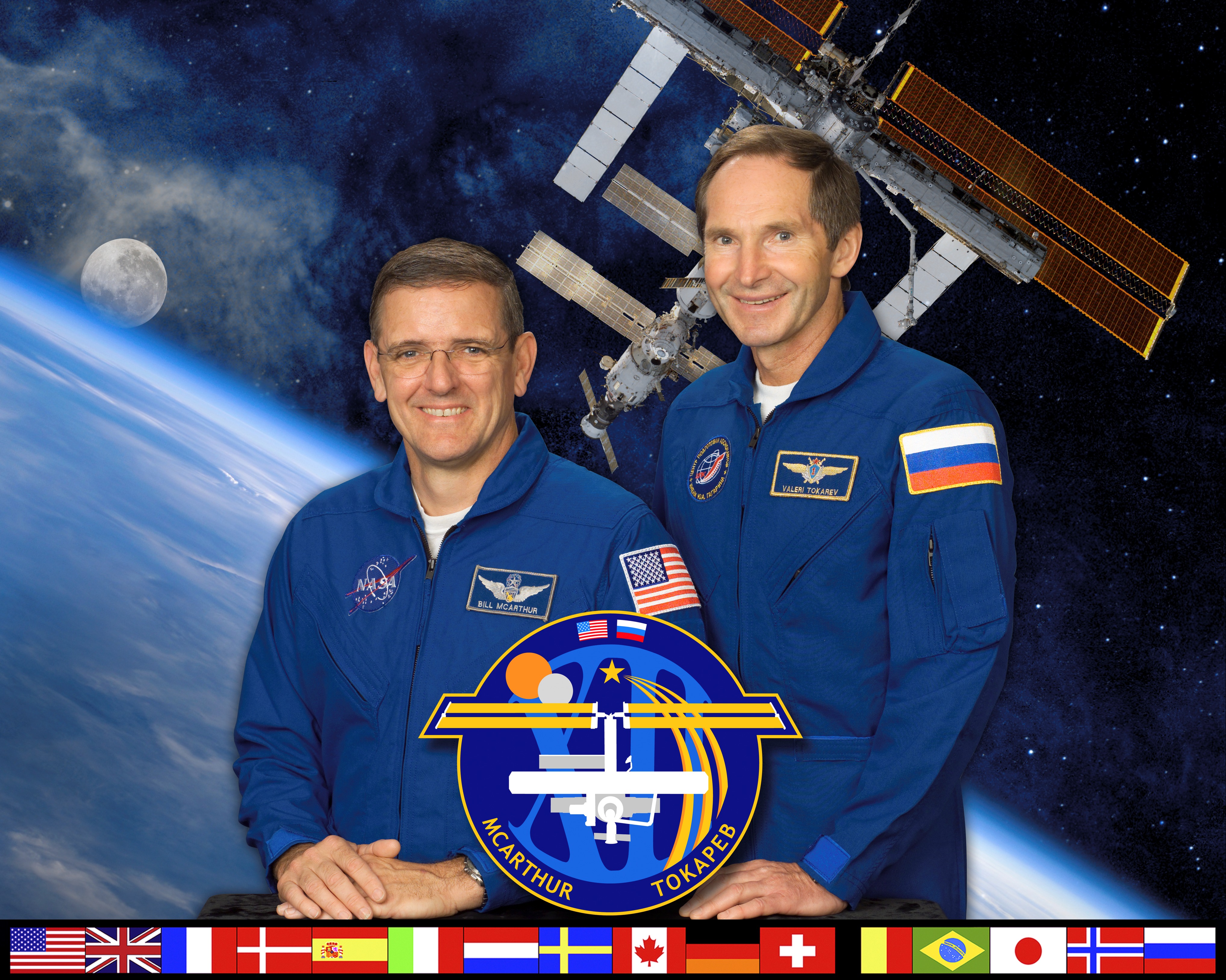 Two male astronauts in blue flight suits pose for their Expedition 12 crew picture with an image of the ISS orbiting the Earth in the background.