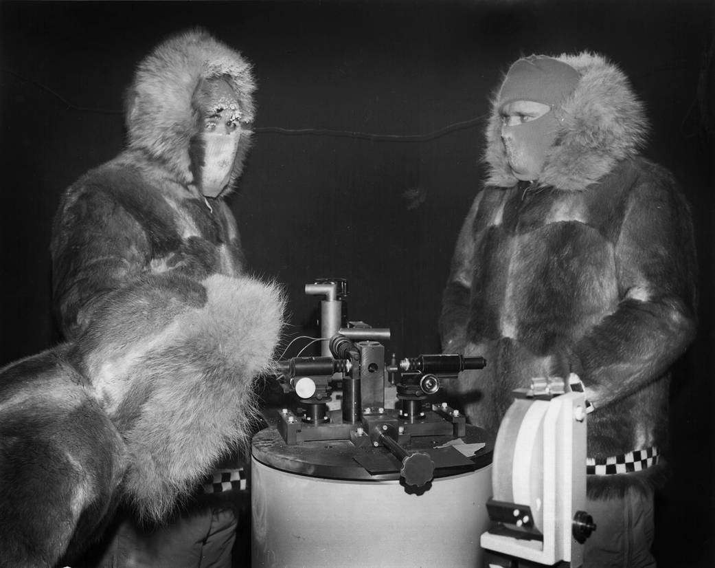 Two men in furs and ski masks work on a piece of hardware