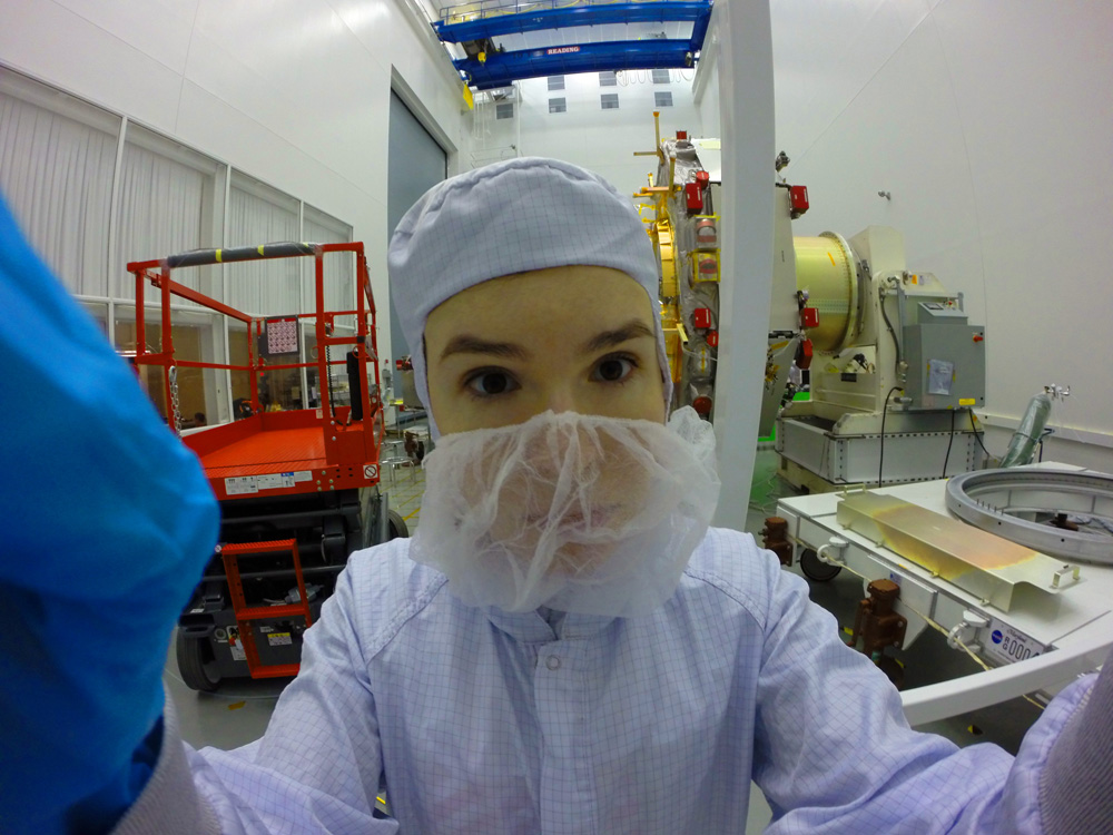 A woman wearing light blue coveralls, blue rubber gloves, and a loose, gauzy mask over her face leans close in a selfie taken with a digital camera. She has dark eyes and her hands appear large because they are closer to the lens. Behind her, a white clean room and red and yellow equipment and lifts are visible.